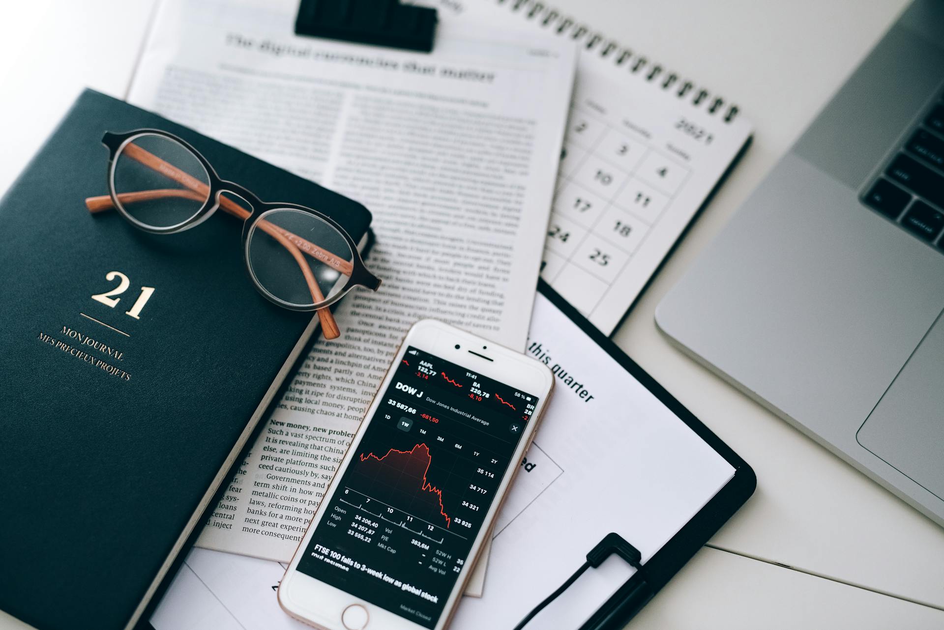 A stylish workspace featuring financial documents, eyeglasses, an iPhone displaying stock data, and a laptop.